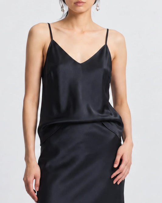 A woman stands against a plain light background wearing the Saint Art Finn Cami Black by Saint Art NY, featuring thin straps and a relaxed fit. Her hands are at her sides, with her face partially visible.