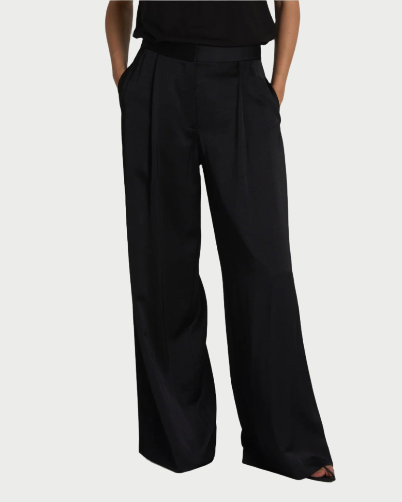 A person presents the Saint Art NY Neve Black Wide Leg Pants, made from luxe charmeuse fabric. These high-waisted, wide-leg pants feature pockets and pleats for a loose fit, styled with a matching black top against a plain backdrop.