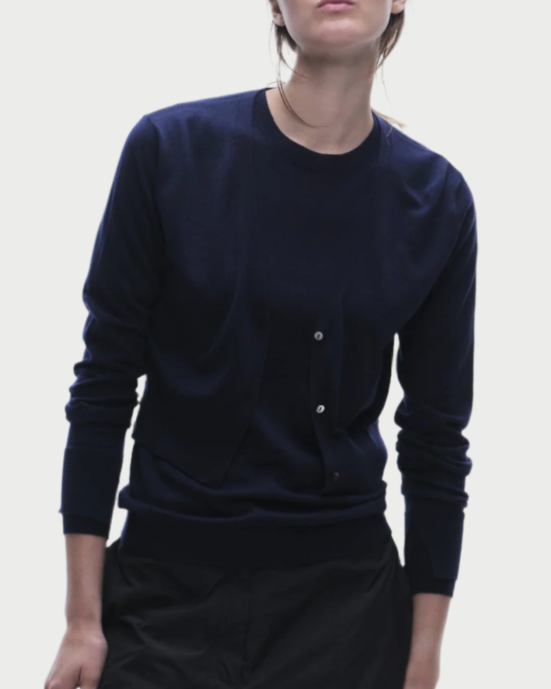 A person wears a navy blue Nixon Cropped Cardigan from Saint Art NY with buttons on the front. They stand against a plain background, one hand in the pocket of dark pants and the other resting at their side, with a slight head tilt.