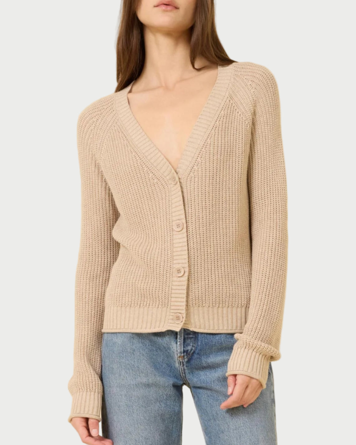 A woman in the One Grey Day Raleigh Cardigan, a beige chunky fisherman-rib knit with buttons and a relaxed fit, pairs it with blue jeans while standing against a plain light background.