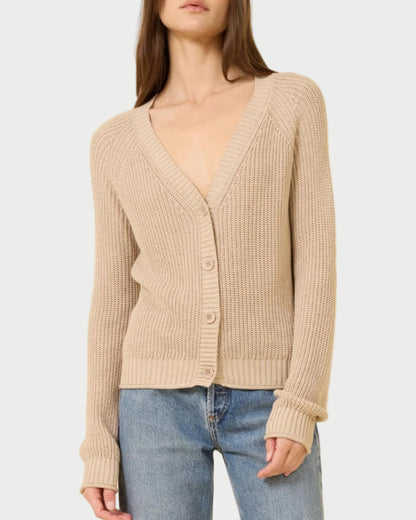 A woman in the One Grey Day Raleigh Cardigan, a beige chunky fisherman-rib knit with buttons and a relaxed fit, pairs it with blue jeans while standing against a plain light background.
