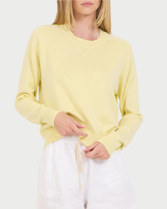 A woman models the Warwick Cotton Pullover by One Grey Day in light yellow, paired with white shorts; she stands against a plain background, with one hand on her waistband and the other holding a drawstring.