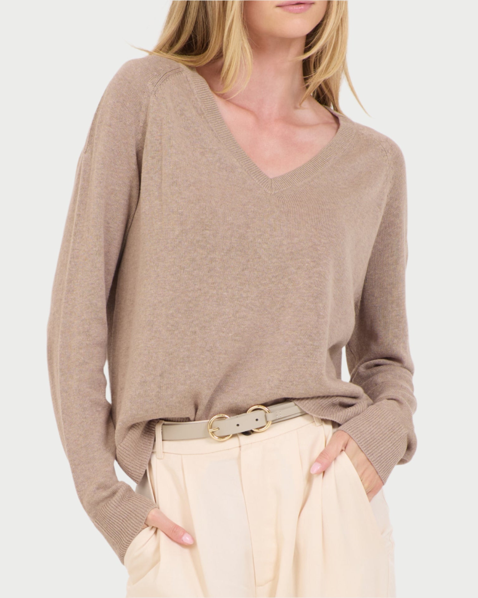 A person in a beige Linen V Neck Sweater by One Grey Day, paired with cream pants and a gold-buckled belt, stands with hands in pockets against a plain light gray background.