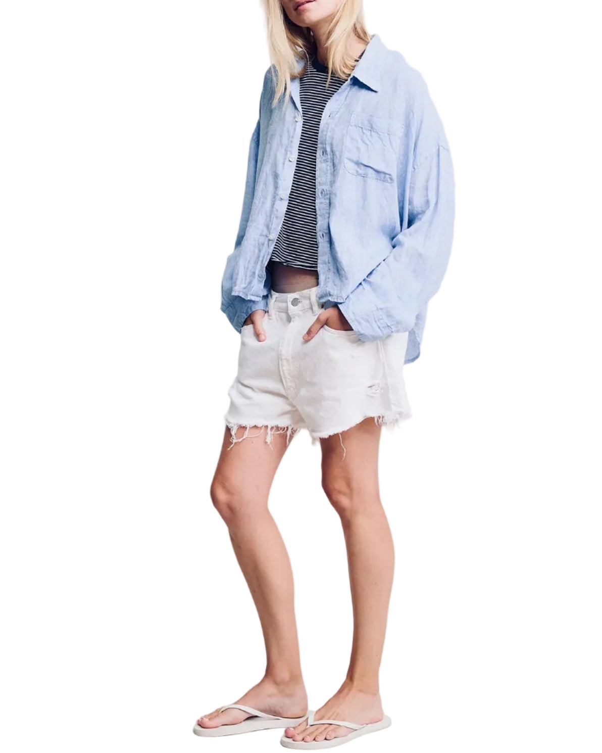 Woman wearing a light blue denim shirt, gray t-shirt, white shorts, and sandals on a plain background