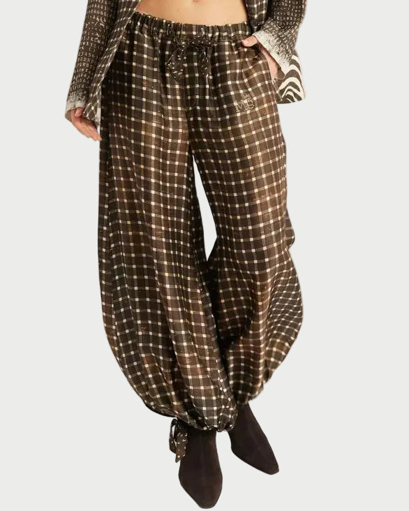 A model stands against a light gray studio background wearing a matching brown and beige checkered blazer and wide-leg harem pants, styled with a black bandeau top and dark brown ankle boots.