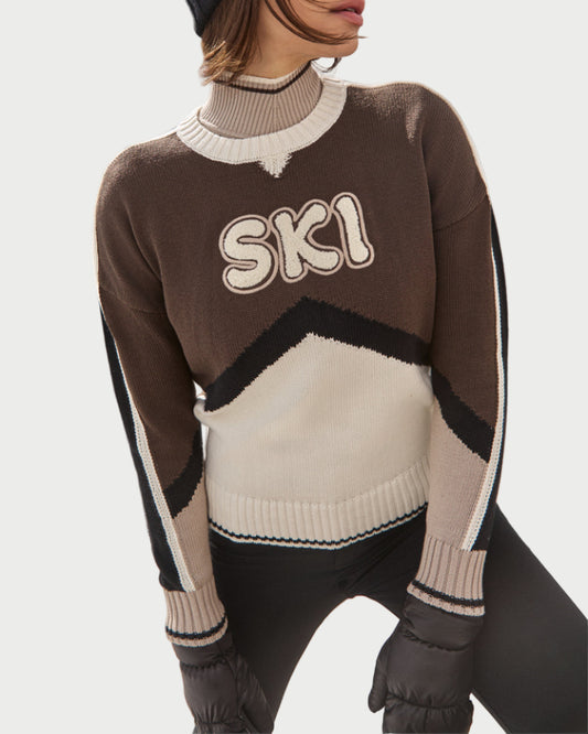 A model wears the Alp N Rock Aiken Ski Sweater in brown, beige, and white with "SKI" on the front, paired with black pants, black gloves, and a black beanie, posing against a plain white background.