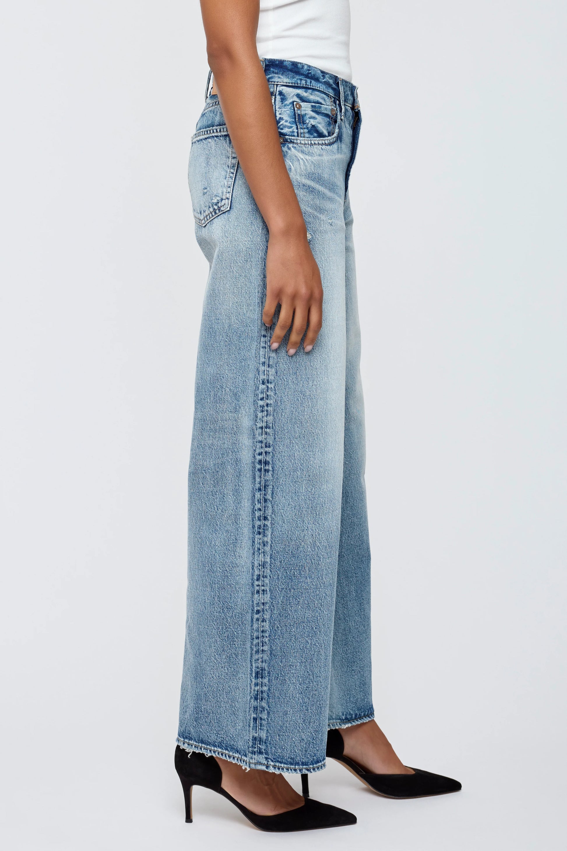 A person stands sideways against a plain light background wearing Moussy Lewes Super Wide Jeans in light blue, paired with a white top and black pointed-toe heels.