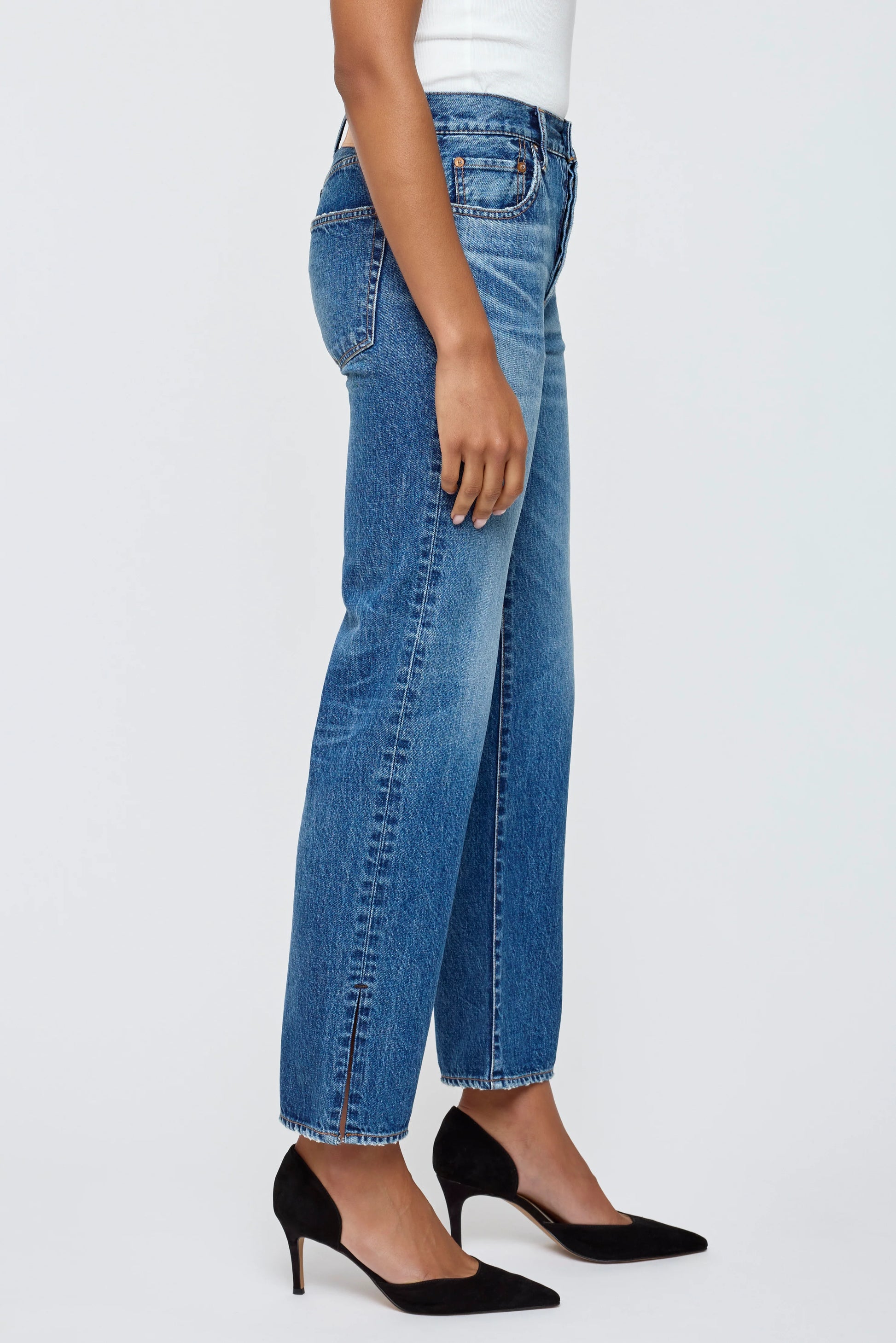A person stands sideways against a plain light background, wearing Moussy Sunset Straight Jeans in Japanese cotton denim, paired with a white sleeveless top and black pointed-toe heels.