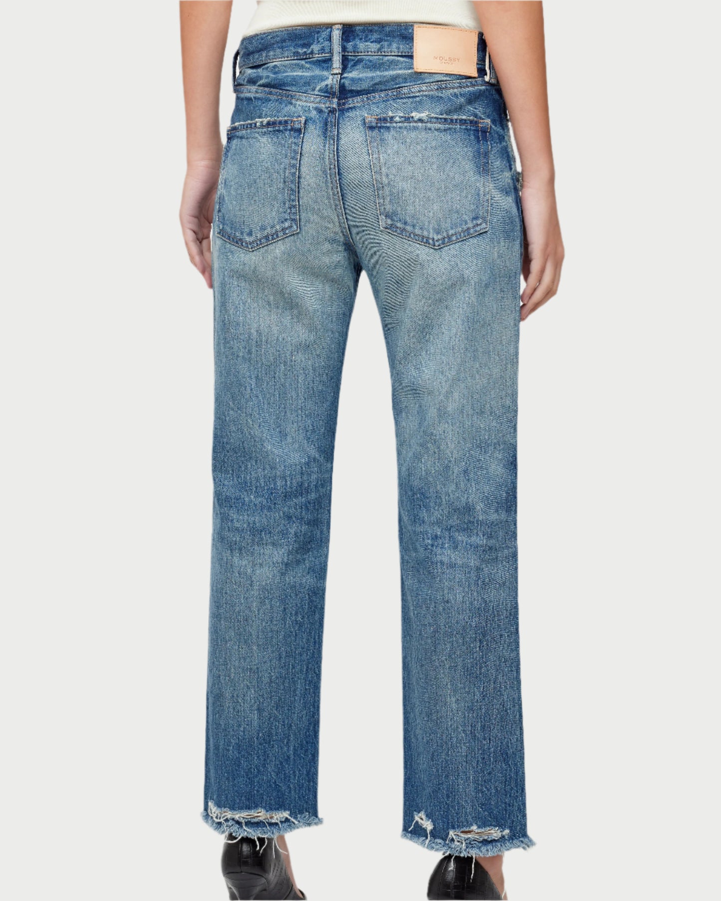 A person is wearing the Sayville Boys Jeans by Moussy, featuring mid-rise, medium-wash, straight-leg style made from Japanese denim with frayed hems and visible back pockets. A patch adorns the waistband. The image shows the jeans from behind against a plain background.