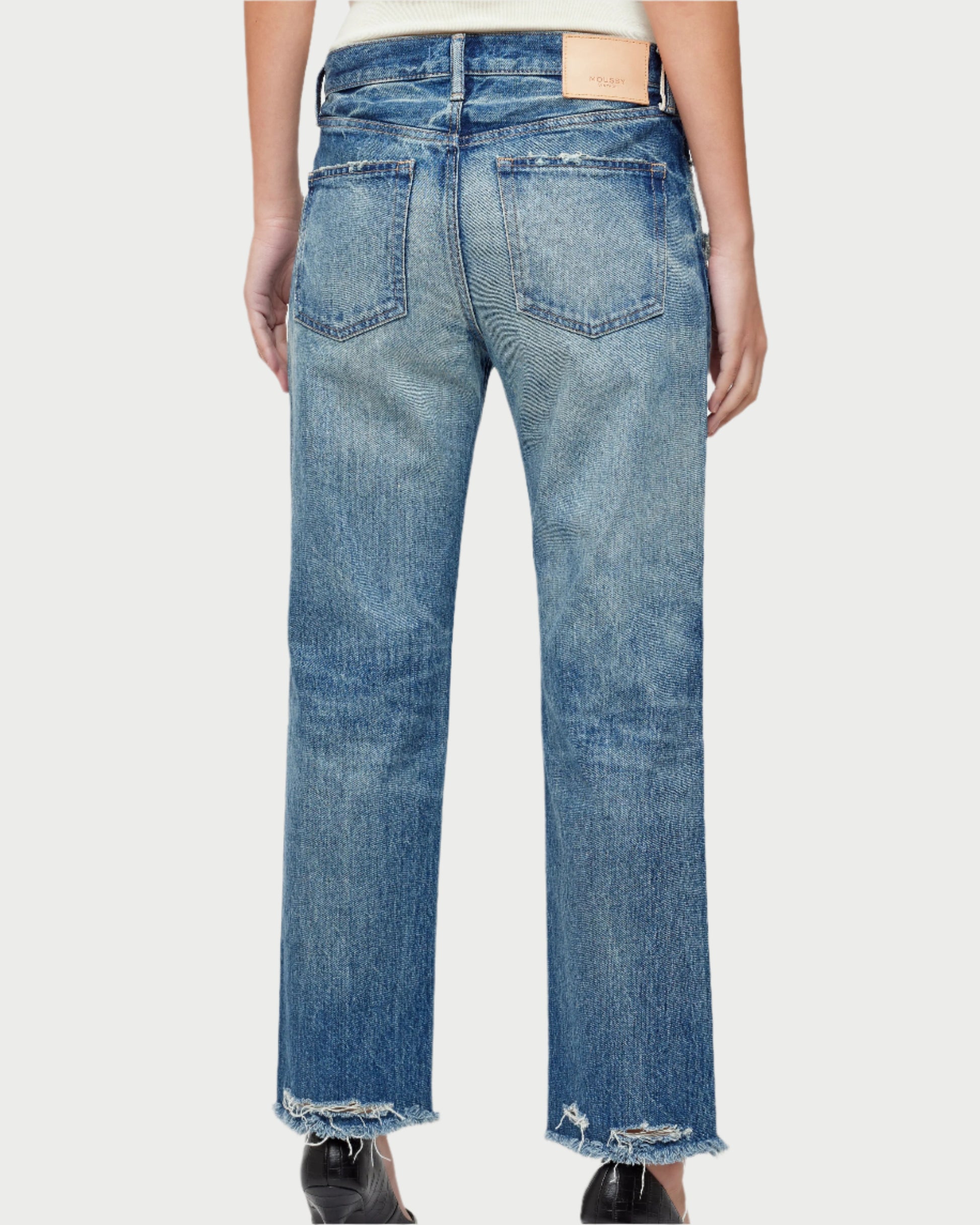 A person is wearing the Sayville Boys Jeans by Moussy, featuring mid-rise, medium-wash, straight-leg style made from Japanese denim with frayed hems and visible back pockets. A patch adorns the waistband. The image shows the jeans from behind against a plain background.