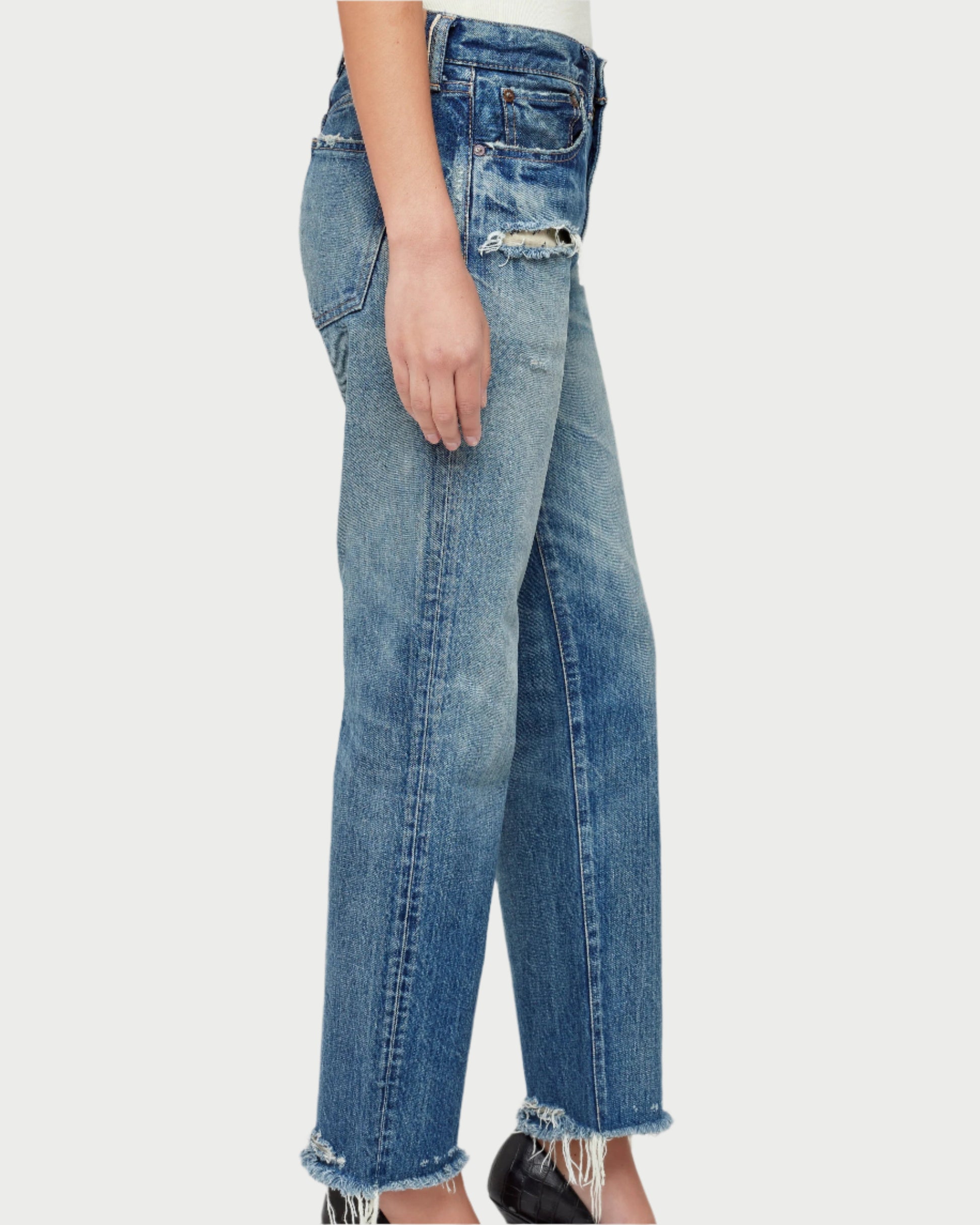 A person stands in profile, showcasing Moussy's Sayville Boys Jeans, crafted from Japanese denim. Featuring frayed hems and torn patches, these mid-rise, boy-fit jeans exude a casual vintage look and are stylishly paired with black shoes for a timeless ensemble.
