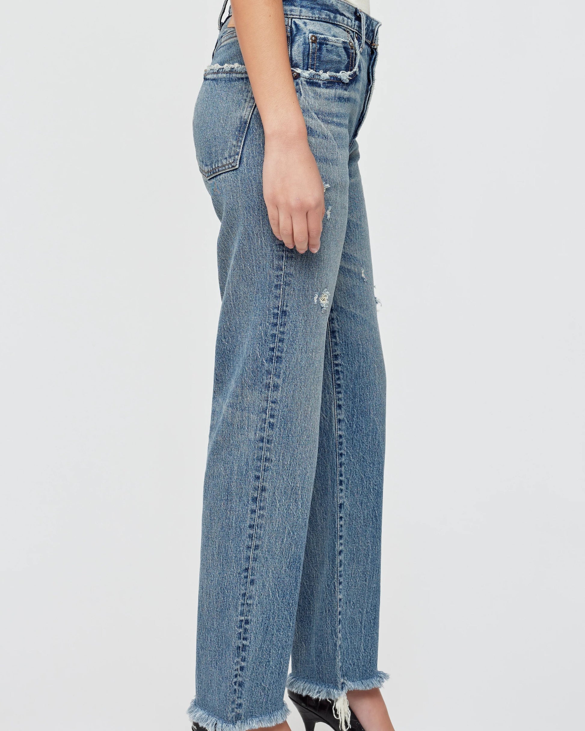 A person stands in profile against a plain light background, wearing Moussy Massapequa Straight Leg Jeans—high-waisted blue jeans crafted from premium Japanese denim, featuring frayed hems and slight distressing.