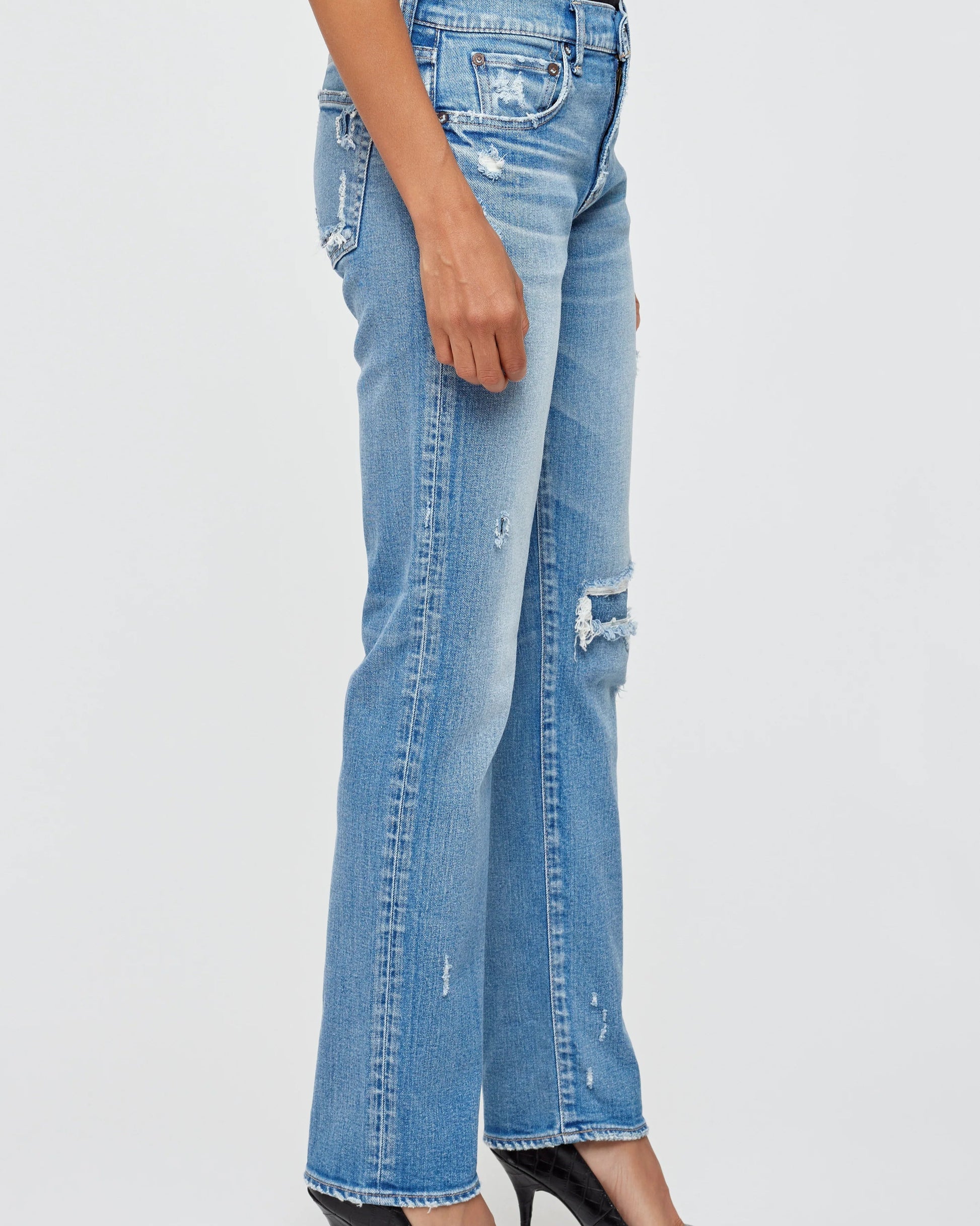 A person stands against a plain light background wearing light blue MOUSSY Fairview Slim Straight Jeans with rips on the thigh and knee, paired with black high-heeled shoes.