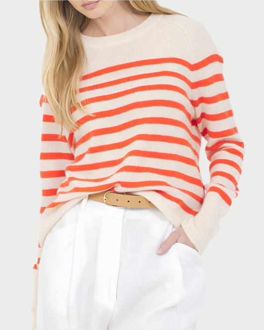 A woman wears the One Grey Day Sloane Cashmere Pullover in beige with bright orange stripes, styled with white high-waisted pants and a tan belt. Her long blonde hair is loose and her hands are in her pockets for a classic look.
