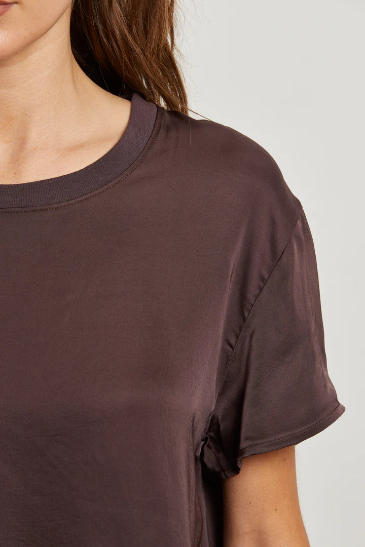 A close-up shows a person in the Delphine Satin Crewneck Tee in Cocoa by perfectwhitetee, featuring rolled cuffs and short sleeves. Only the left side of their upper body and part of their face appear against a plain background.