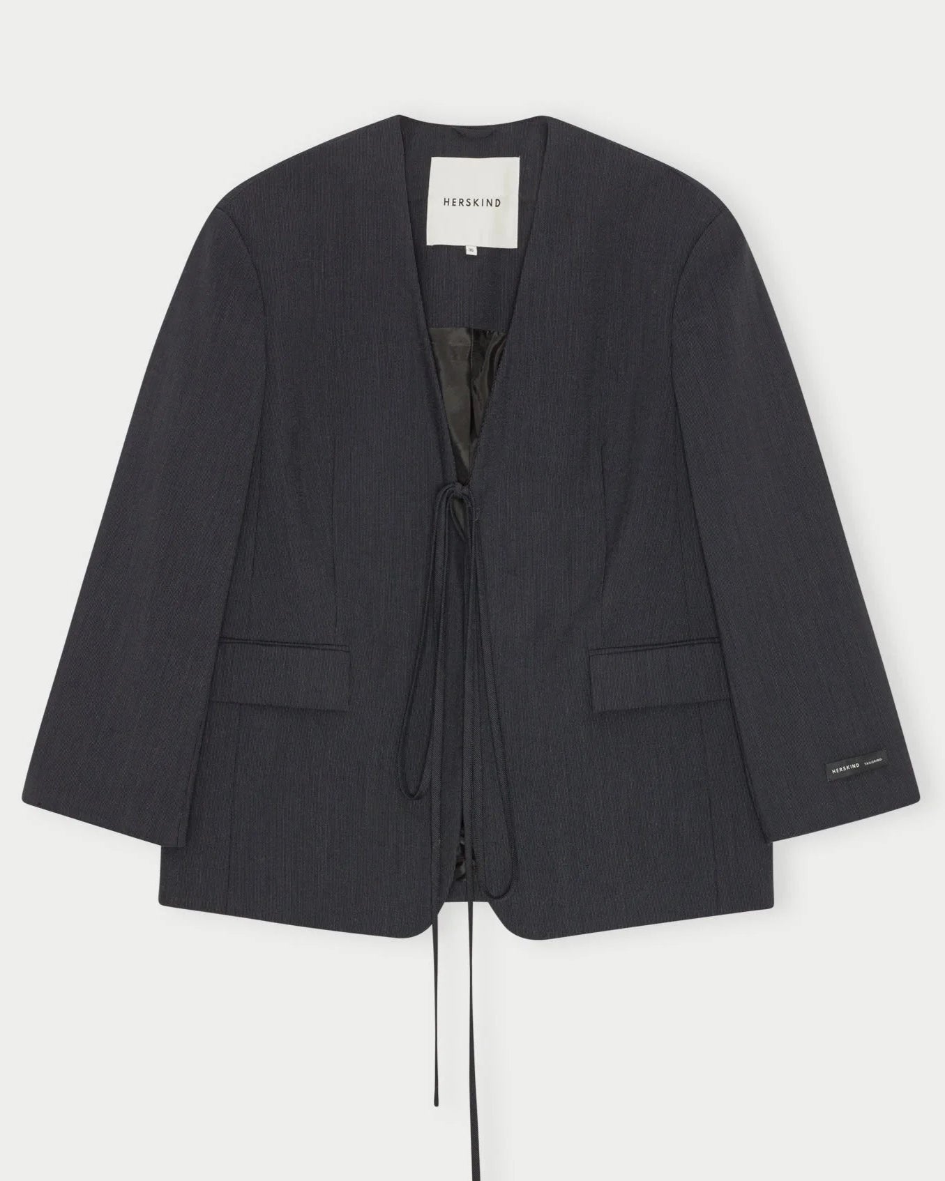 The Indiana Blazer by Herskind is a collarless, oversized blazer in dark charcoal with a deep V-neck, two front pockets, and front tie closure. Crafted from premium navy melange wool with a minimalist design. Inside label reads "HERSKIND.