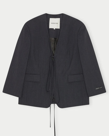 The Indiana Blazer by Herskind is a collarless, oversized blazer in dark charcoal with a deep V-neck, two front pockets, and front tie closure. Crafted from premium navy melange wool with a minimalist design. Inside label reads "HERSKIND.