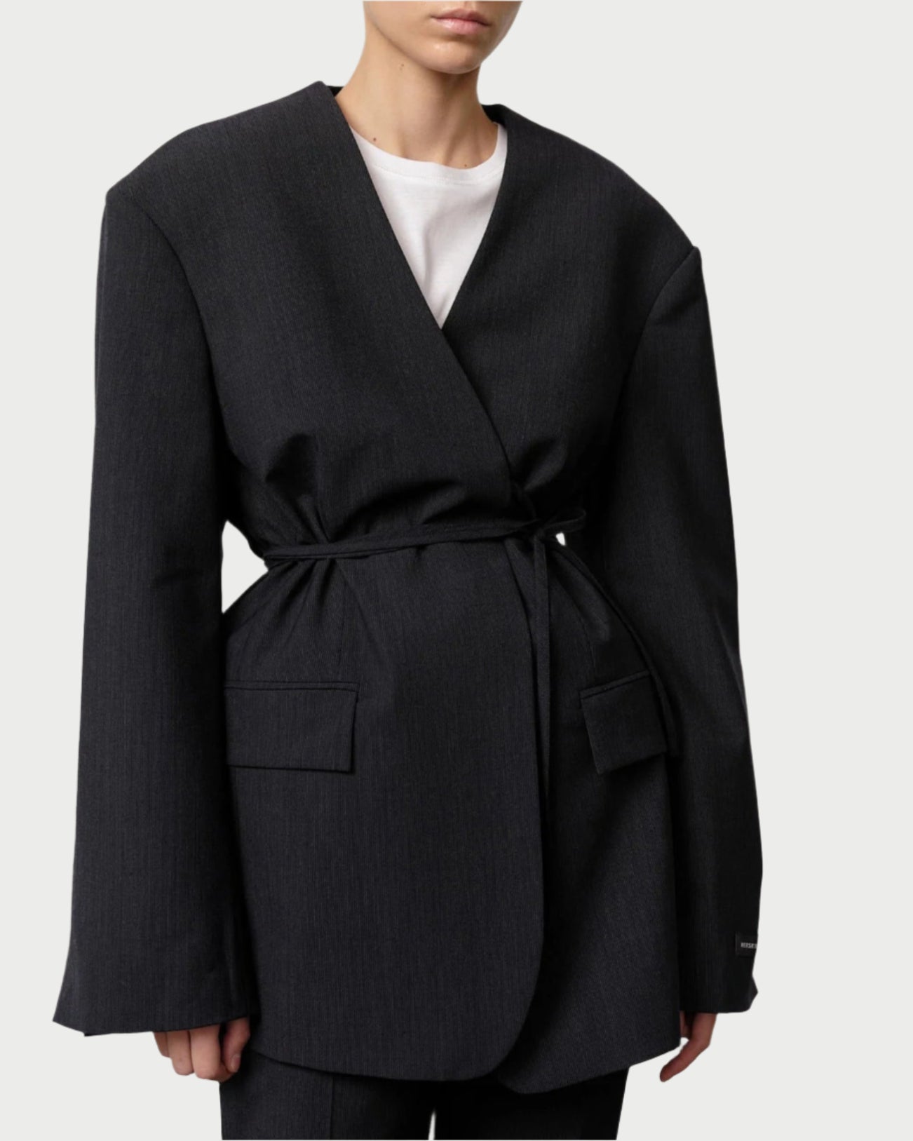 A person wears the Herskind Indiana Blazer in oversized navy melange wool with wide shoulders, wrap-style front, tie belt waist, large front pockets, and a white shirt underneath; their head is partially cropped out.