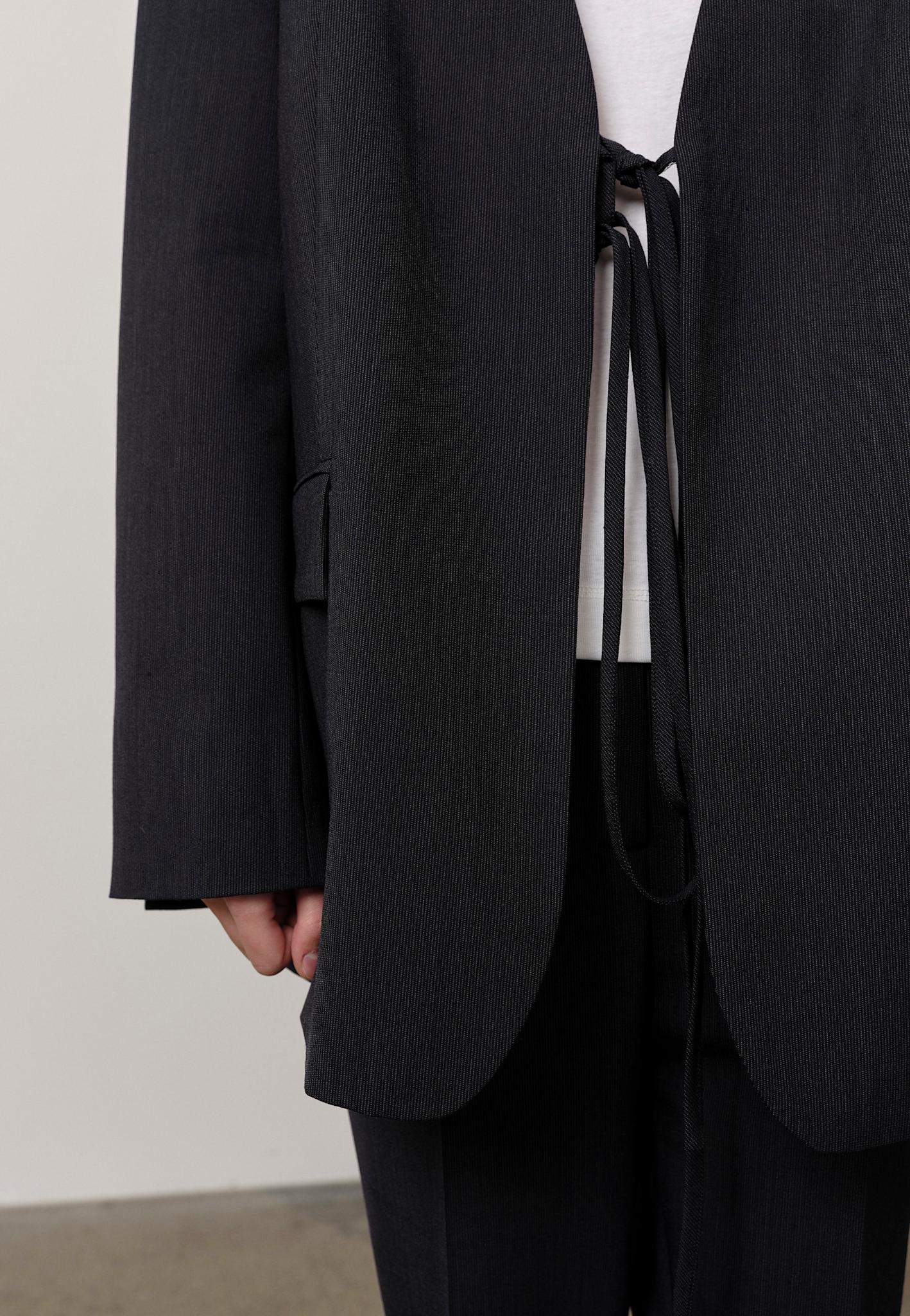 A person wears the Herskind Indiana Blazer—an oversized black pinstripe suit with a relaxed fit and tie closure—over a white shirt. Only the lower torso, one hand, and upper legs are visible.