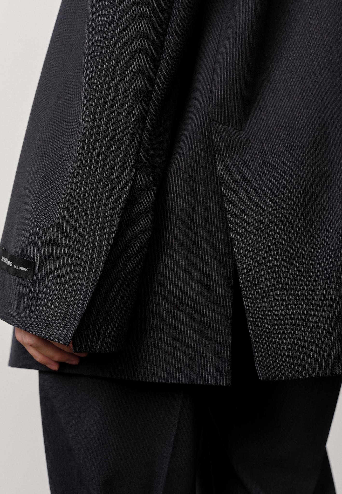 A close-up shows a person wearing the Herskind Indiana Blazer in dark pinstripe, with a small black label on the cuff. Their hand is partially visible by their side against a plain, light background.