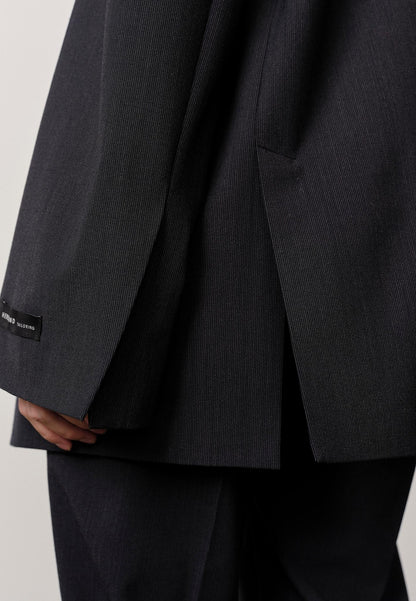 A close-up shows a person wearing the Herskind Indiana Blazer in dark pinstripe, with a small black label on the cuff. Their hand is partially visible by their side against a plain, light background.