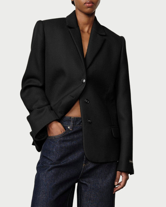 Person wearing a black blazer and dark jeans against a plain background
