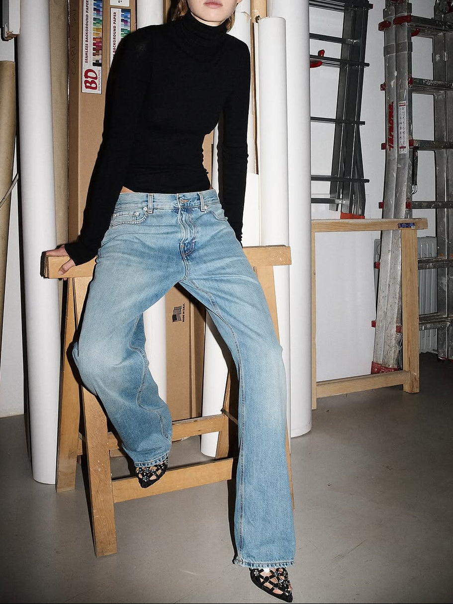 Person wearing a black turtleneck and blue jeans sitting on a wooden stool in an industrial setting.