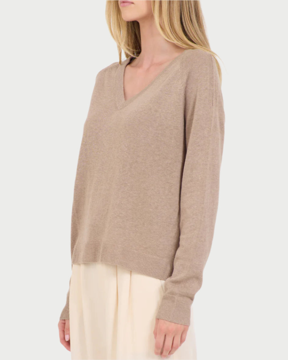 A woman stands against a plain white background, wearing a One Grey Day Linen V Neck Sweater made from a linen/cotton blend, elegantly paired with a light-colored skirt.