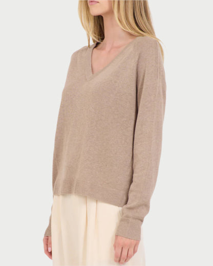 A woman stands against a plain white background, wearing a One Grey Day Linen V Neck Sweater made from a linen/cotton blend, elegantly paired with a light-colored skirt.