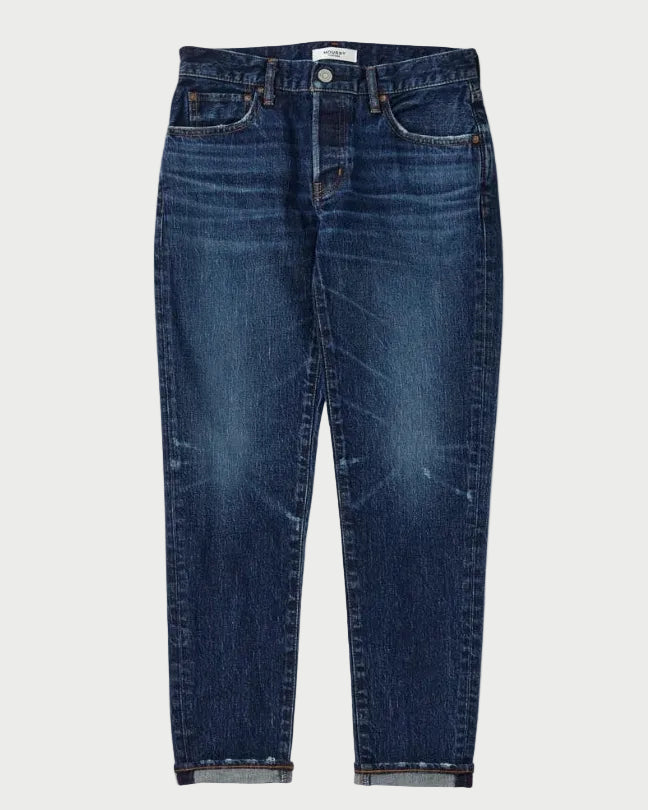 A pair of dark blue denim jeans with a tapered, cropped fit and rolled cuffs, shown flat against a light gray background.