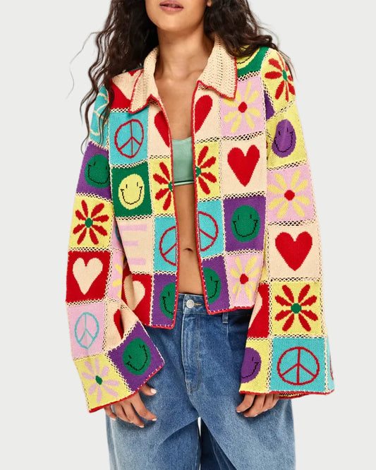 A woman wears the ME369 Miami Crochet Cardigan featuring colorful granny-square patchwork with hearts, peace signs, smiley faces, and flowers. The cardigan has wide sleeves and a collar, styled with a green top and blue jeans.