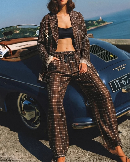A woman wearing a brown and beige plaid two-piece lounge set with a black bralette, posing next to a classic blue Porsche convertible outdoors by the sea.