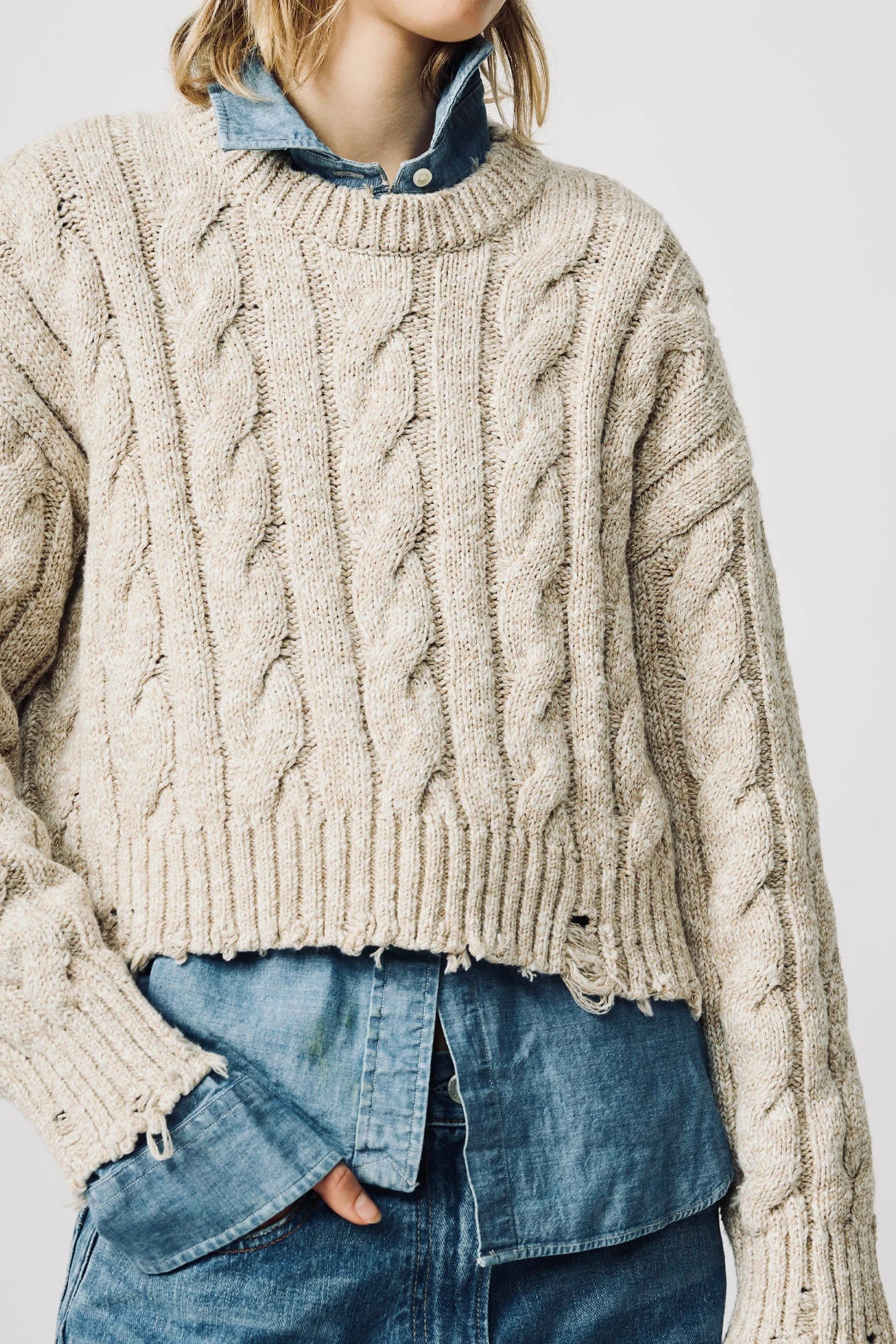 A close-up of a person wearing a chunky cable-knit sweater in a warm oatmeal beige color, layered over a blue denim shirt and paired with blue jeans.