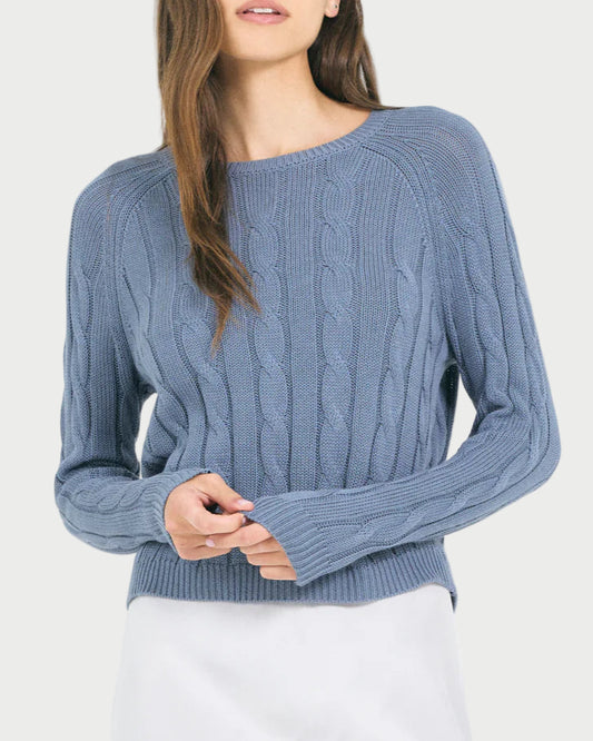 A woman stands against a plain light background, wearing the One Grey Day Raleigh Cable Pullover in light blue with long sleeves and a white skirt. Her hair partly covers her face as she gently holds the sweater sleeve.