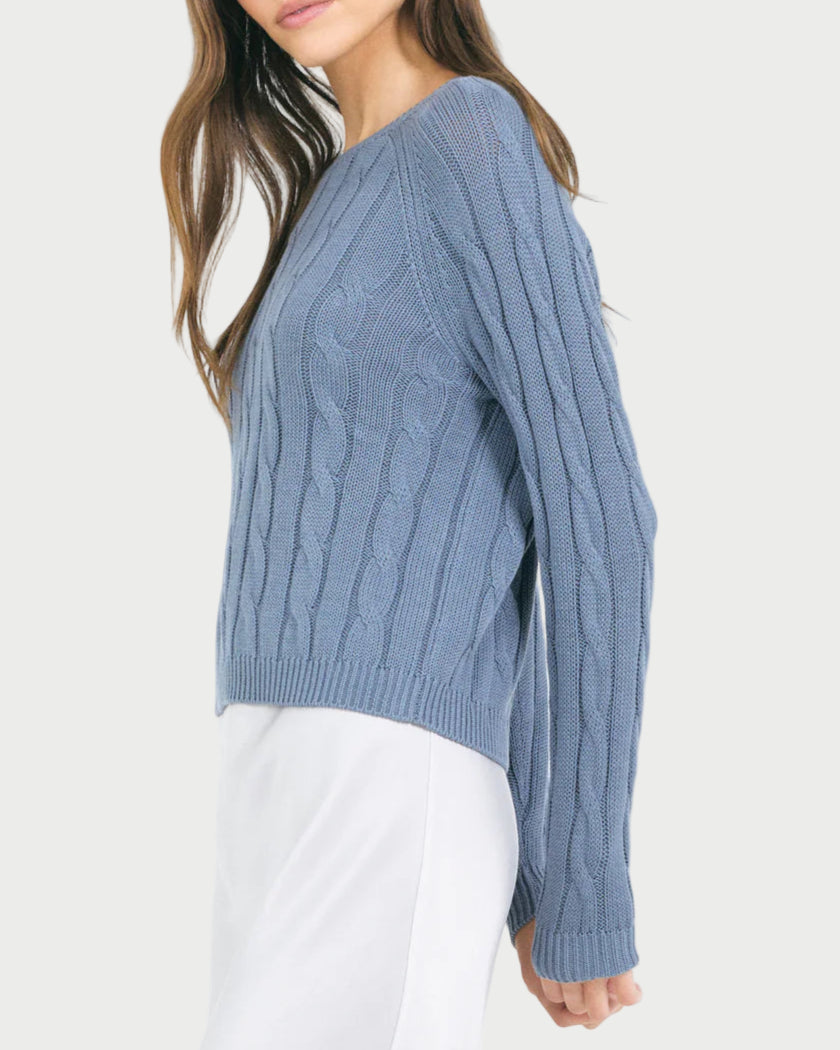 A woman stands against a plain light background in a light blue, long-sleeve One Grey Day Raleigh Cable Pullover with a crew neck, paired with a white skirt.