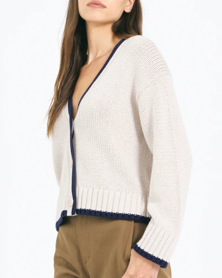A woman wears the Mallory Cardigan by One Grey Day, a cream V-neck cardigan with navy trim, relaxed fit, and button front, paired with brown pants. Her face is partially out of frame.