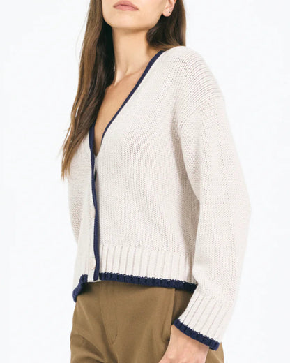 A woman wears the Mallory Cardigan by One Grey Day, a cream V-neck cardigan with navy trim, relaxed fit, and button front, paired with brown pants. Her face is partially out of frame.