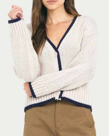 A woman wears the Mallory Cardigan by One Grey Day—a cream, relaxed-fit knit cardigan with navy trim—paired with brown pants, posing with one arm crossed and the other hand touching her hair.