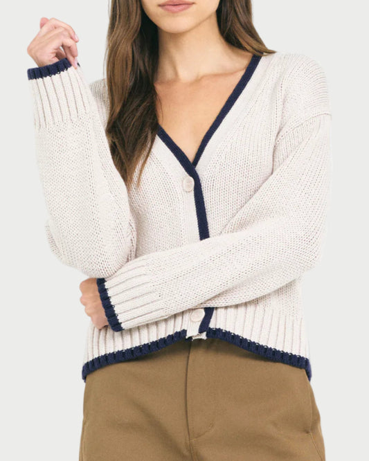 A woman wears the Mallory Cardigan by One Grey Day—a cream, relaxed-fit knit cardigan with navy trim—paired with brown pants, posing with one arm crossed and the other hand touching her hair.
