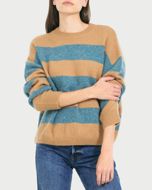 A person in the One Grey Day Sophie Pullover—a relaxed fit striped sweater with wide tan and teal stripes by One Grey Day—and blue jeans stands with one arm crossed against a plain light background.