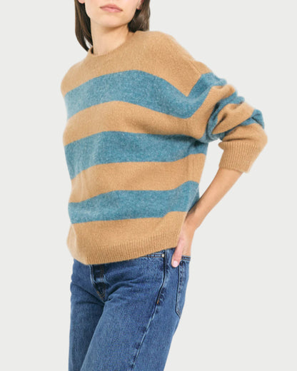 A person wearing the One Grey Day Sophie Pullover—a relaxed fit, tan and blue striped merino cashmere sweater with long sleeves—paired with blue jeans, stands with one hand on their hip against a plain background.