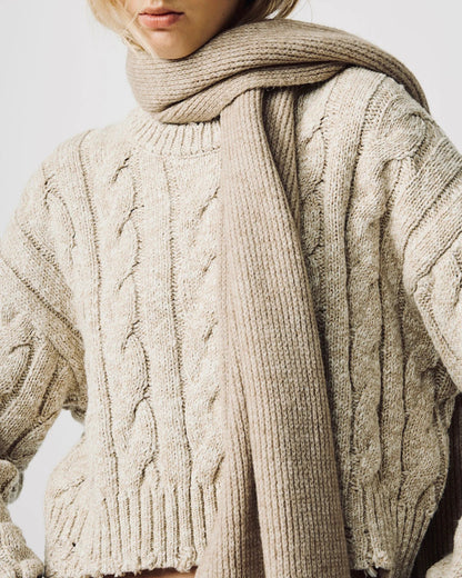 A close-up of a woman wearing a chunky cable-knit sweater and a matching thick knit scarf in a warm oatmeal beige color, set against a plain light gray background.