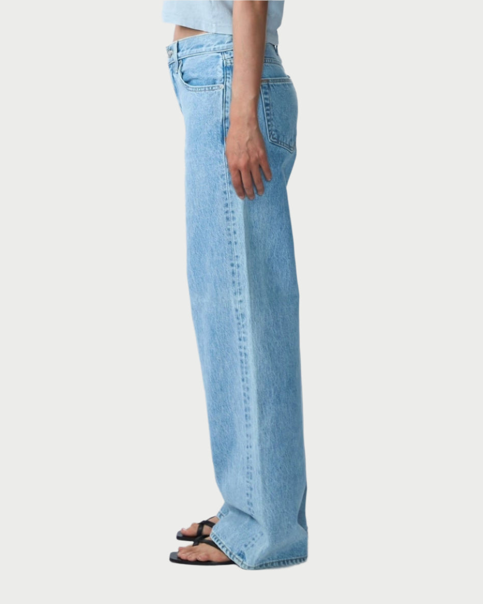 Side profile of a person with long dark hair, wearing a light blue t-shirt and SLVRLAKE's Mica Jeans in Clear Skies, paired with black sandals against a stark white background.