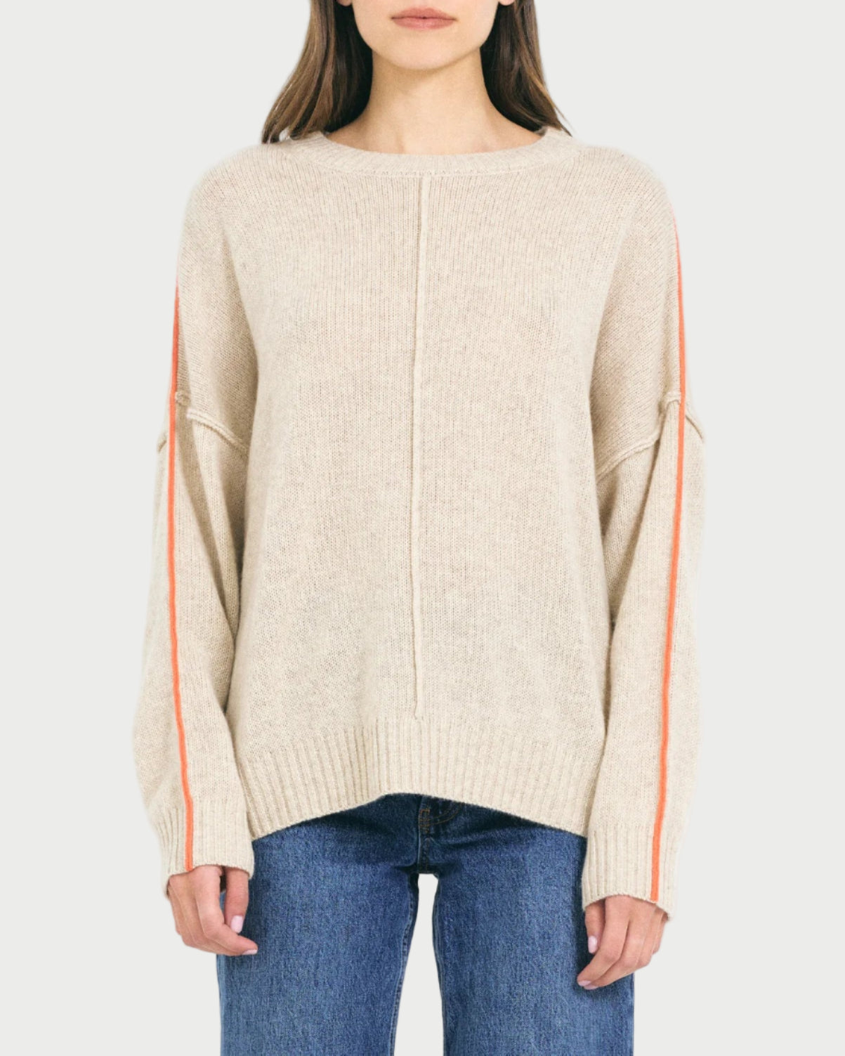 A woman wears the One Grey Day Greta Cashmere Crewneck, an oversized beige sweater with orange sleeve stripes from One Grey Day, paired with blue jeans and standing against a plain light background.