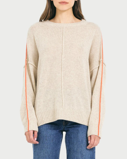 A woman wears the One Grey Day Greta Cashmere Crewneck, an oversized beige sweater with orange sleeve stripes from One Grey Day, paired with blue jeans and standing against a plain light background.