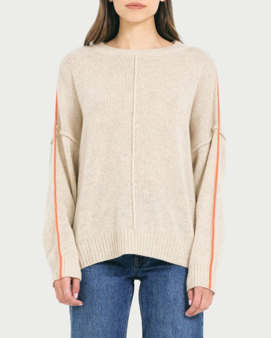 A woman wears the One Grey Day Greta Cashmere Crewneck, an oversized beige sweater with orange sleeve stripes from One Grey Day, paired with blue jeans and standing against a plain light background.