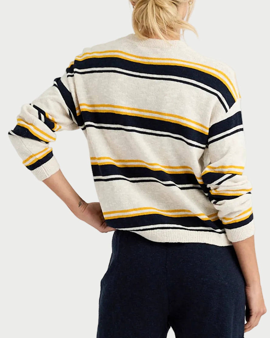 A person with blonde hair in a ponytail, seen from behind, wears the One Grey Day Susannah Pullover—a cream sweater with navy and yellow stripes—and dark pants, resting their left hand on their hip.