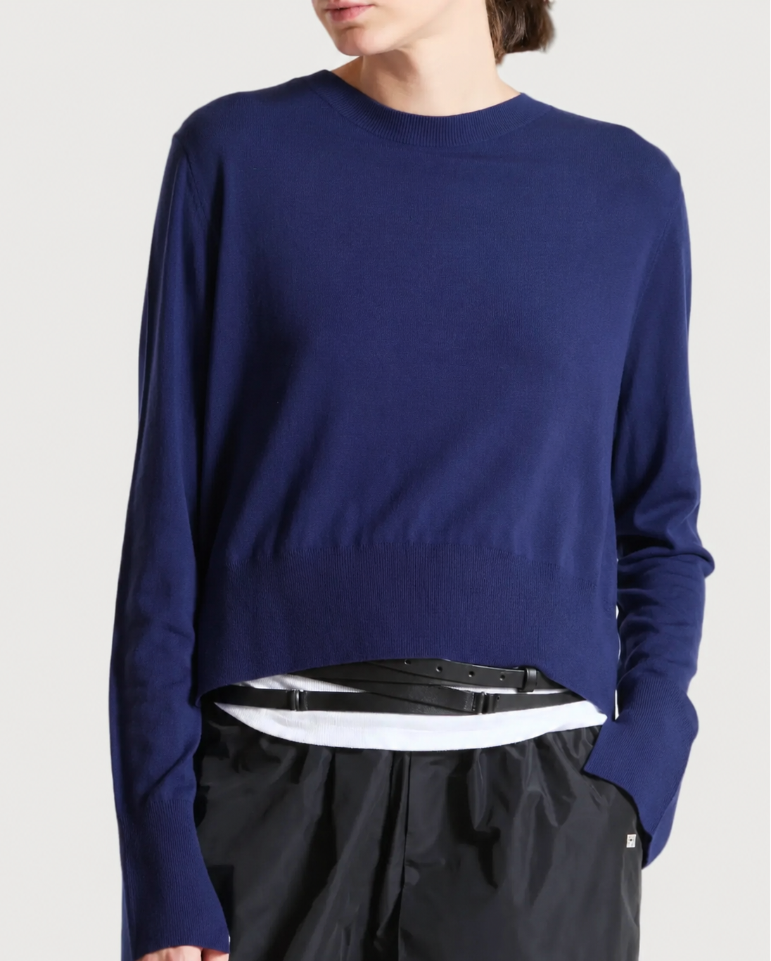 A person wearing a navy blue long-sleeve crewneck sweater over a white shirt and black pants, shown from the neck down against a light gray studio background.