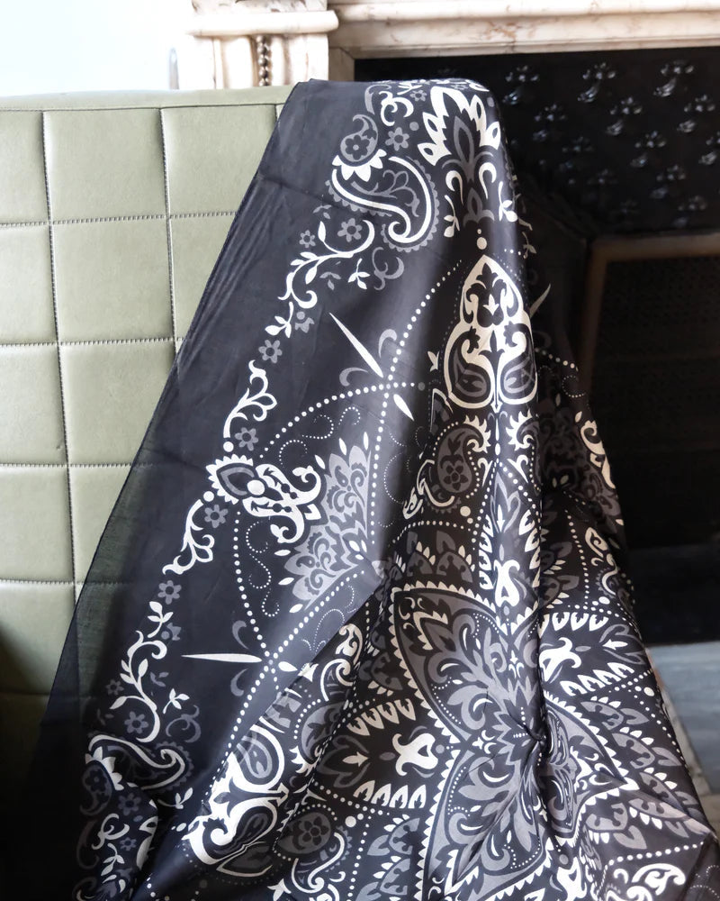 A Maison Lecomte Flament Bandana Scarf Black is draped over the back of a green, quilted chair beside a stone or marble fireplace.