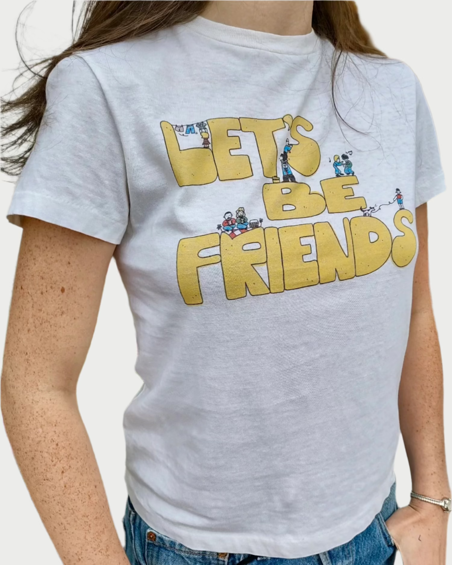 A person wearing the RE/DONE Lets Be Friends Classic Tee, featuring playful yellow text "LET'S BE FRIENDS" adorned with small cartoon characters engaged in various activities on the front. The versatile white t-shirt adds charm to their look as they stand outdoors in front of a potted plant with pink flowers.